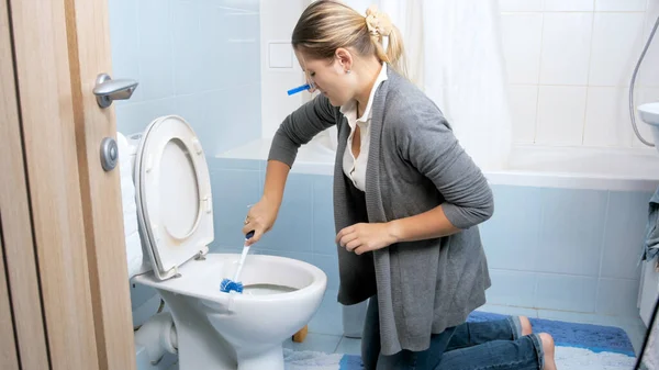Mujer joven cerrando la nariz con pinza de ropa mientras lava y limpia el inodoro — Foto de Stock