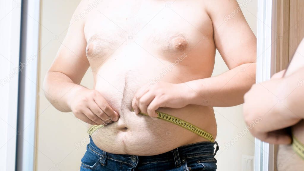Yougn fat man with big belly stuggling to measure his size. Concept of male overweight, weight loss and dieting