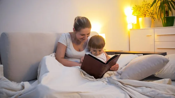 Молодая улыбающаяся мать с маленьким ребенком сын читает книгу в постели — стоковое фото