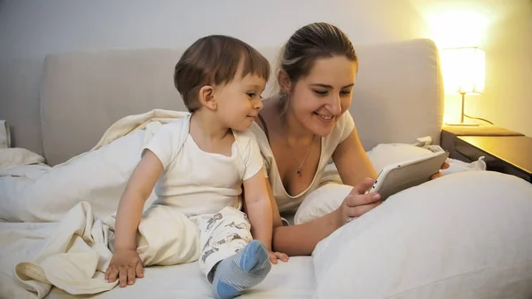 Feliz madre sonriente con niño pequeño viendo vídeo en la tableta por la noche —  Fotos de Stock