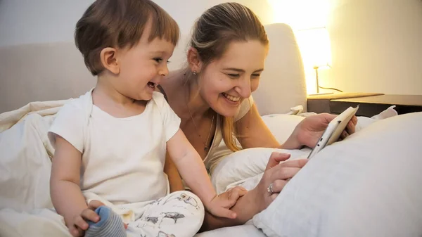 Portret van vrolijke lachende moeder met peuter zoon liggend in bed 's nachts en met behulp van tablet computer — Stockfoto