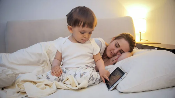 Cute little boy using tablet computer while mother is sleeping in bed — Stock Photo, Image
