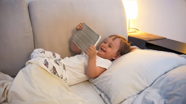 Glad skrattande småbarn pojke liggande i sängen på natten och titta på tecknad film på digital surfplatta dator — Stockfoto