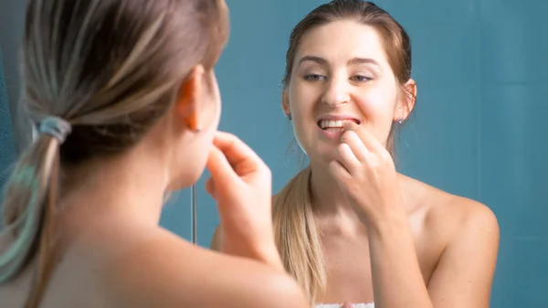 Jonge vrouw schoonmaken en controleren van haar tanden bij spiegel in badkamer — Stockfoto