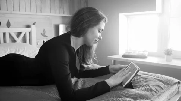 Черно-белый портрет улыбающейся деловой женщины, лежащей на кровати и работающей на цифровом планшетном компьютере — стоковое фото