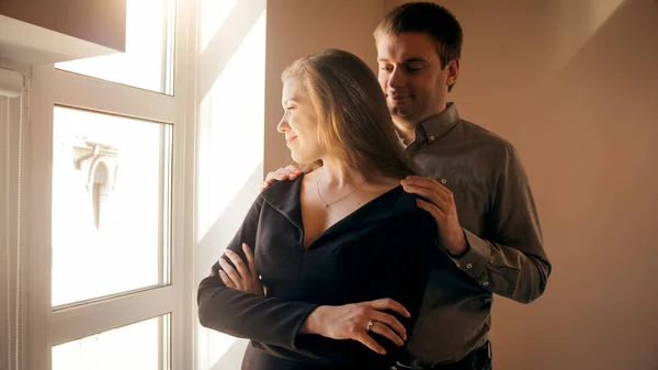 Ung kärleksfull man kramar sin fru tittar ut genom fönstret på solnedgången — Stockfoto