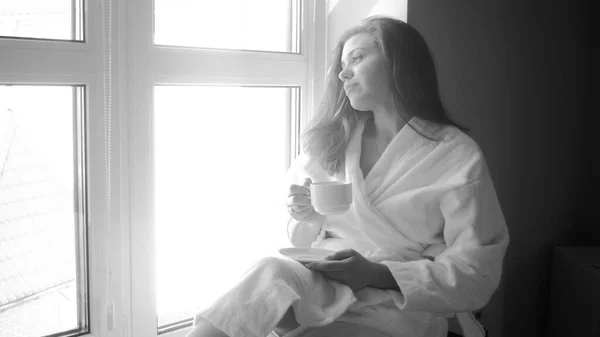 Retrato en blanco y negro de una joven en albornoz sentada en una ventana grande y tomando café —  Fotos de Stock