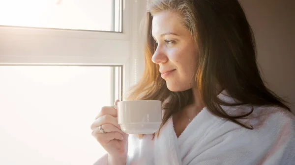 Porträtt av leende kvinna dricka kaffe och titta ut genom fönstret på morgonen — Stockfoto