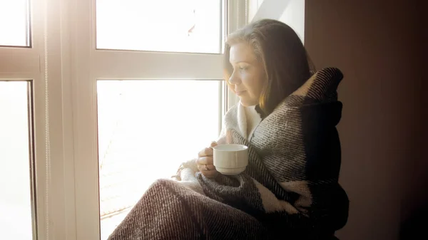 Mladá žena covereng v kostkovaném sedí u okna a pije čaj — Stock fotografie