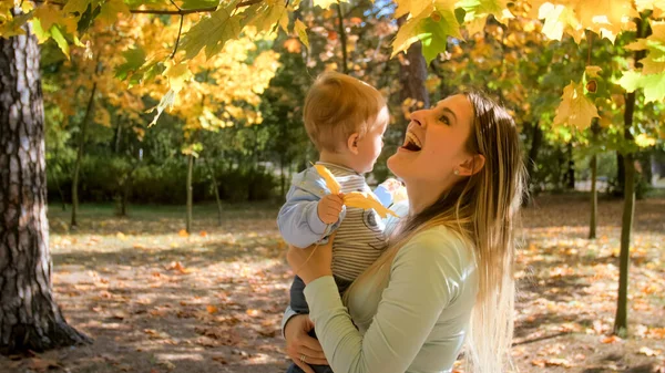 Portræt af glad smilende mor, der holder og krammer hendes lille søn, der ser på efterårsbladene i parken - Stock-foto