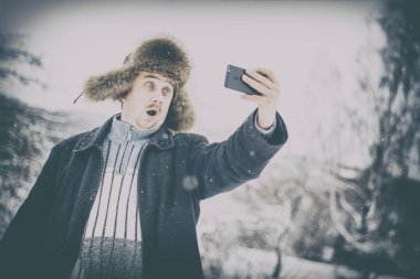Genç bir adam kameraya şaşırır, karlı kış ormanda bir selfie yapar. Sokaktaki adam. üzerinde Doğal kürk şapka ürünleri. Orada tonlama.