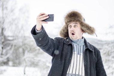 Genç bir adam kameraya şaşırır, karlı kış ormanda bir selfie yapar. Sokaktaki adam. üzerinde Doğal kürk şapka ürünleri. Orada tonlama.