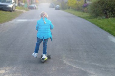 Arkadan bak. Scooter süren mavi ceketli bir kız. Sarı saç rengi. Ayağını tahtaya yuvarlıyor..