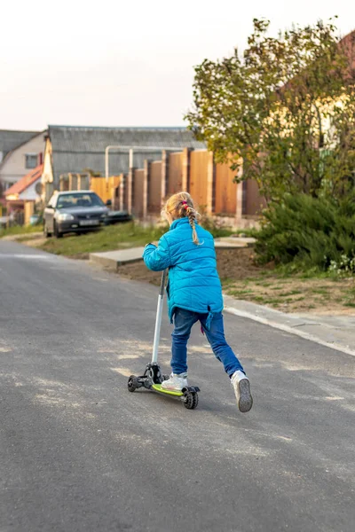 Vista para trás. menina em um casaco azul montando uma scooter. cor de cabelo loiro. ela rola o pé no tabuleiro . — Fotografia de Stock