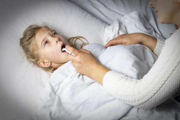 Seorang gadis dengan rambut putih berbaring di tempat tidur. ibu menggunakan inhaler membuat suntikan di tenggorokan pasien . — Stok Foto