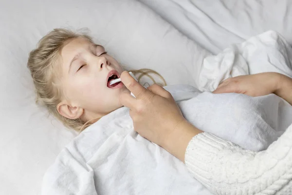 Seorang gadis dengan rambut putih berbaring di tempat tidur. ibu menggunakan inhaler membuat suntikan di tenggorokan pasien . — Stok Foto