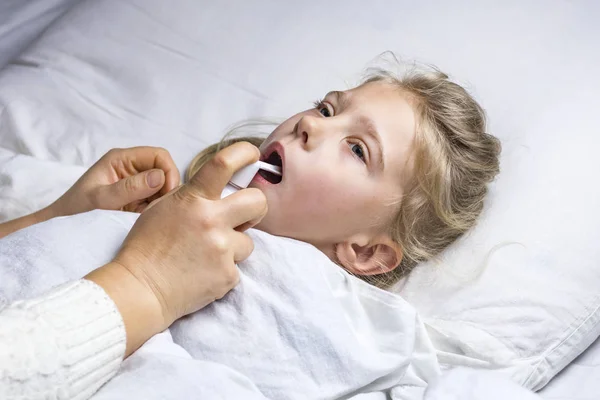 Ένα κορίτσι με άσπρα μαλλιά ξαπλώνει στο κρεβάτι. Η μαμά που χρησιμοποιεί έναν εισπνευστήρα κάνει μια ένεση στο λαιμό ενός ασθενούς. — Φωτογραφία Αρχείου