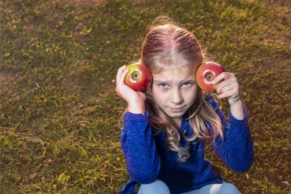 Yumuşak odaklanma. Gün ışığı. Beyaz saçlı bir kız elinde iki kırmızı elma tutuyor. Güneş batıyor.