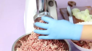 Elektrikli beyaz et öğütücüsü. Kıyma dilimlenmiş eti metal bir kaseye koyar. Mavi eldivenli bir insan eli her şeyi düzeltir.