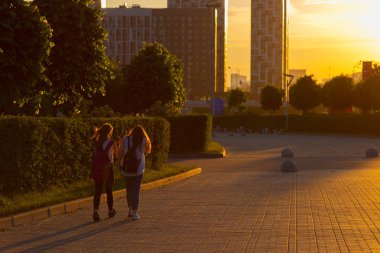 Arka ışıklı siluetleri. Yüzler görünür değil. Fayans ve yeşil çalılar ve ağaçlar ile döşeli sokak boyunca günbatımı yürüyüş adlı iki kız. Sıcak bahar romantik akşam. Arka plan vasıl modern mimari yapı