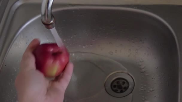 男はこすり 流水の下で熟した赤いリンゴを洗います ベジタリアンのスナックを準備します 水の流れの下で台所の流しの上の果物と左の人間の手 上からの眺め ステンレス調理器具 — ストック動画