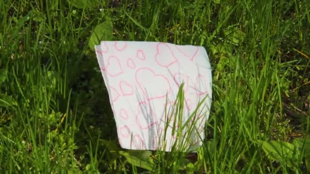 Serviette en papier blanc avec des contours de coeur rouge repose sur l'herbe verte — Video