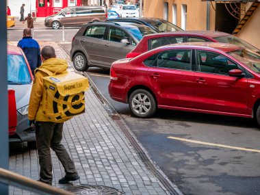 Moskova, Rusya - 14 Eylül 2019: Park halindeki araçların yanında sarı Yandex-Eda sırt çantası ile markalı üniformalı yemek dağıtım hizmeti nin kuryesi. Yandex Gıda sembolleri ve logosu ile termal çanta