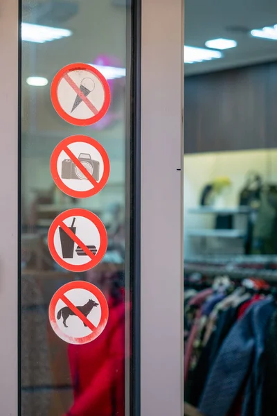 Запретные знаки у входа в магазин одежды. Наклейки на стеклянном окне возле двери с запретами. Красными кругами не допускаются значки с символами - фотоаппарат, еда, мороженое, собака — стоковое фото
