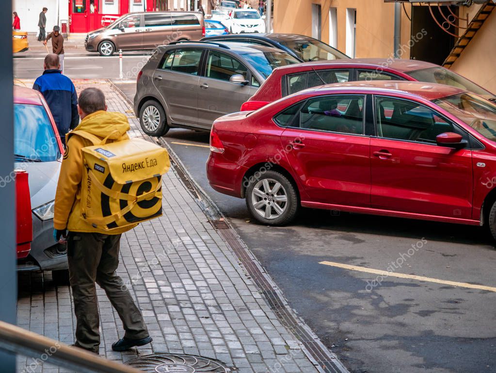 Moscow, Russia - September 14, 2019: Courier of food delivery service in a branded uniform with a yellow Yandex-Eda backpack next to parked cars. Thermal bag with symbols and logo of Yandex Food