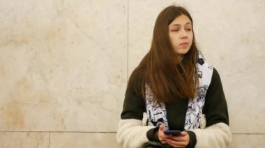 Genç esmer kadın metroda akıllı telefonlu bir bankta birini bekliyor.