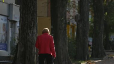 Kırmızı ceketli yaşlı bir kadın sonbaharda gün batımında kaldırımda yürür.