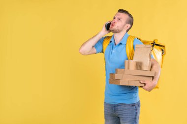 Teslimatçı adam elinde yiyecek kutularıyla telefonda müşteriyle konuşuyor. Yüzüne yemek kuryesi kızgın. Mavi üniformalı ve sırtında buzdolabı çantası var.