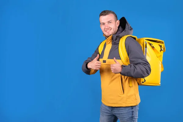 Young smiling caucasian food delivery guy in yellow uniform and refrigerator bag on his back waves finger up in approval or husky on blue background. Courier delivers groceries home.