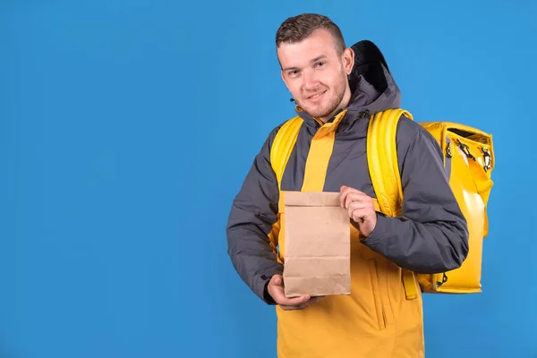 Sırtında sarı üniformalı ve serin çantalı beyaz yemek dağıtım elemanı mavi stüdyo arka planında müşteri için hazır bekleyen kağıt el çantası tutuyor. Kurye ürünleri eve getiriyor.