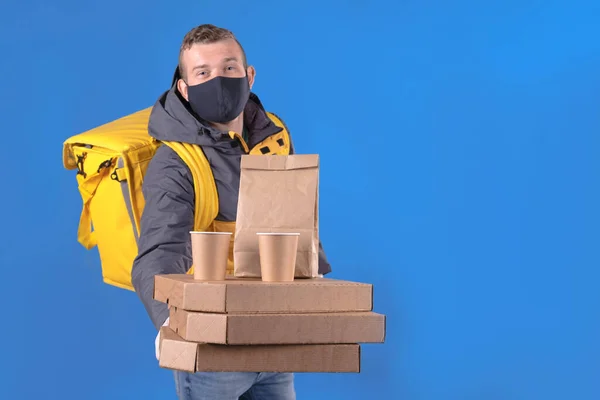 Young caucasian food delivery man in protective black mask and gloves against virus and bacteria is dressed in yellow uniform, cooler bag on his back holds an order on blue fone. Safe food delivery