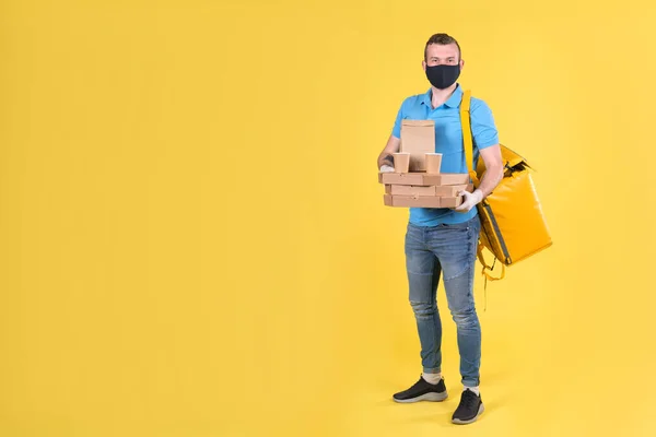 Young food delivery guy in protective mask and gloves is holding an order from restaurant for customer, dressed in blue Polo shirt and carrying yellow shopping bag on his shoulders. Safe food delivery