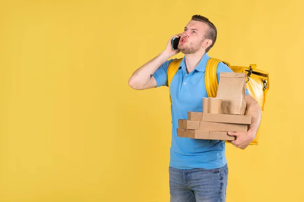 El repartidor sostiene cajas de comida en sus manos y habla emocionalmente por teléfono con el cliente, enojado en la cara, el mensajero de comida está vestido con uniforme azul y bolsa de refrigerador en su espalda en amarillo. — Foto de Stock