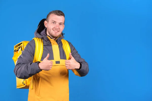 Young smiling caucasian food delivery guy in yellow uniform and refrigerator bag on his back waves finger up in approval or husky on blue background. Courier delivers groceries home.