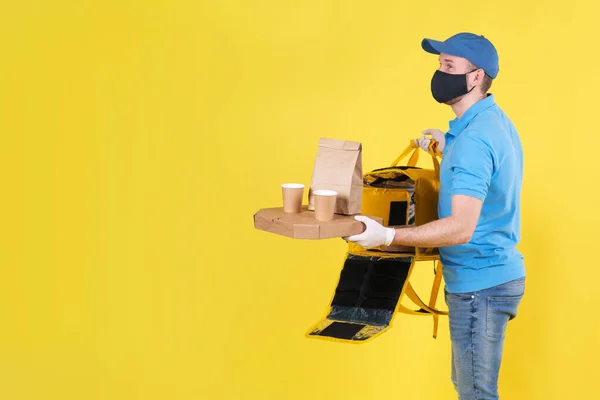 Courier wearing virus protection mask and medical food delivery gloves with yellow open thermos bag holds out an order from restaurant for customer on yellow studio background. safe delivery of food — Stock Photo, Image