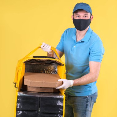 Virüs koruma maskesi takan kurye ve sarı termos çantalı tıbbi gıda dağıtım eldivenleri sarı stüdyo arka planında müşteri için restorandan sipariş veriyor. Güvenli yiyecek teslimatı.