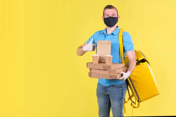 Young smiling food delivery guy in protective mask and gloves in blue uniform holds an order from restaurant for pizza and coffee and waves finger up. safe food delivery to home or office in epidemic.
