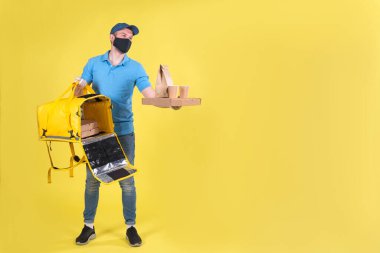 Virüs koruma maskesi takan kurye ve sarı termos çantalı tıbbi gıda dağıtım eldivenleri sarı stüdyo arka planında müşteri için restorandan sipariş veriyor. Güvenli yiyecek teslimatı.