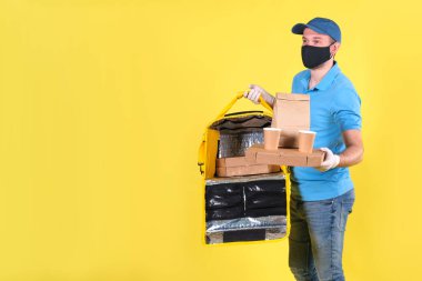 Virüs koruma maskesi takan kurye ve sarı termos çantalı tıbbi gıda dağıtım eldivenleri sarı stüdyo arka planında müşteri için restorandan sipariş veriyor. Güvenli yiyecek teslimatı.