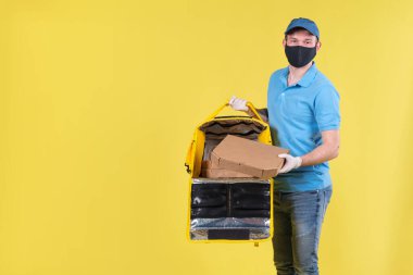 Virüs koruma maskesi takan kurye ve sarı termos çantalı tıbbi gıda dağıtım eldivenleri sarı stüdyo arka planında müşteri için restorandan sipariş veriyor. Güvenli yiyecek teslimatı.