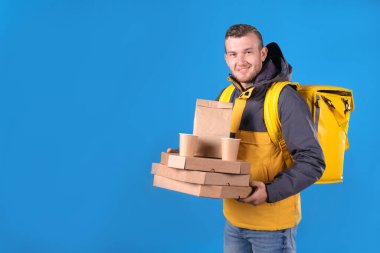 Sarı üniformalı, elinde pizza kutuları, kese kağıdında yemek ve termoçanta mavisi arka planda gülümseyen traşsız kurye portresi. Hızlı, lezzetli ve kaliteli yemek teslimatı kavramı