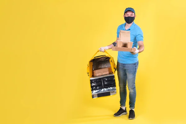 Virüs koruma maskesi takan kurye ve sarı termos çantalı tıbbi gıda dağıtım eldivenleri sarı stüdyo arka planında müşteri için restorandan sipariş veriyor. Güvenli yiyecek teslimatı.