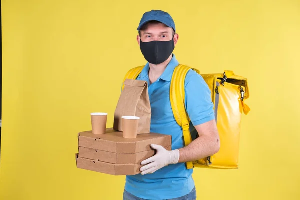 Young food delivery guy in protective mask and gloves is holding an order from restaurant for customer, dressed in blue Polo shirt and carrying yellow shopping bag on his shoulders. Safe delivery.