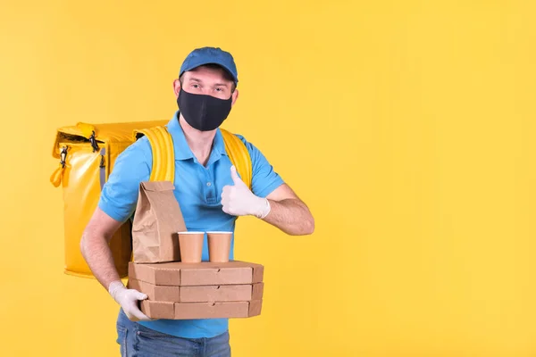 Young smiling food delivery guy in protective mask and gloves in blue uniform holds an order from restaurant for pizza and coffee and waves finger up. safe food delivery to home or office in epidemic.
