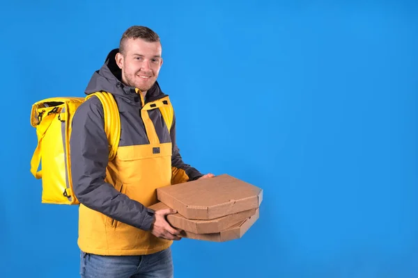 Happy food delivery man holds pizzas in cardboard boxes and holds them out for customer, dressed in yellow uniform and refrigerator bag on his back on blue background. Courier food delivery to home.