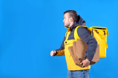 Tıraşsız, beyaz, genç, sarışın adam, markalı sarı üniformalı yemek teslimatçısı elinde kutularca pizzayla restorandan geç saatte mavi arka planda çekilen müşteriye teslim ediyor..